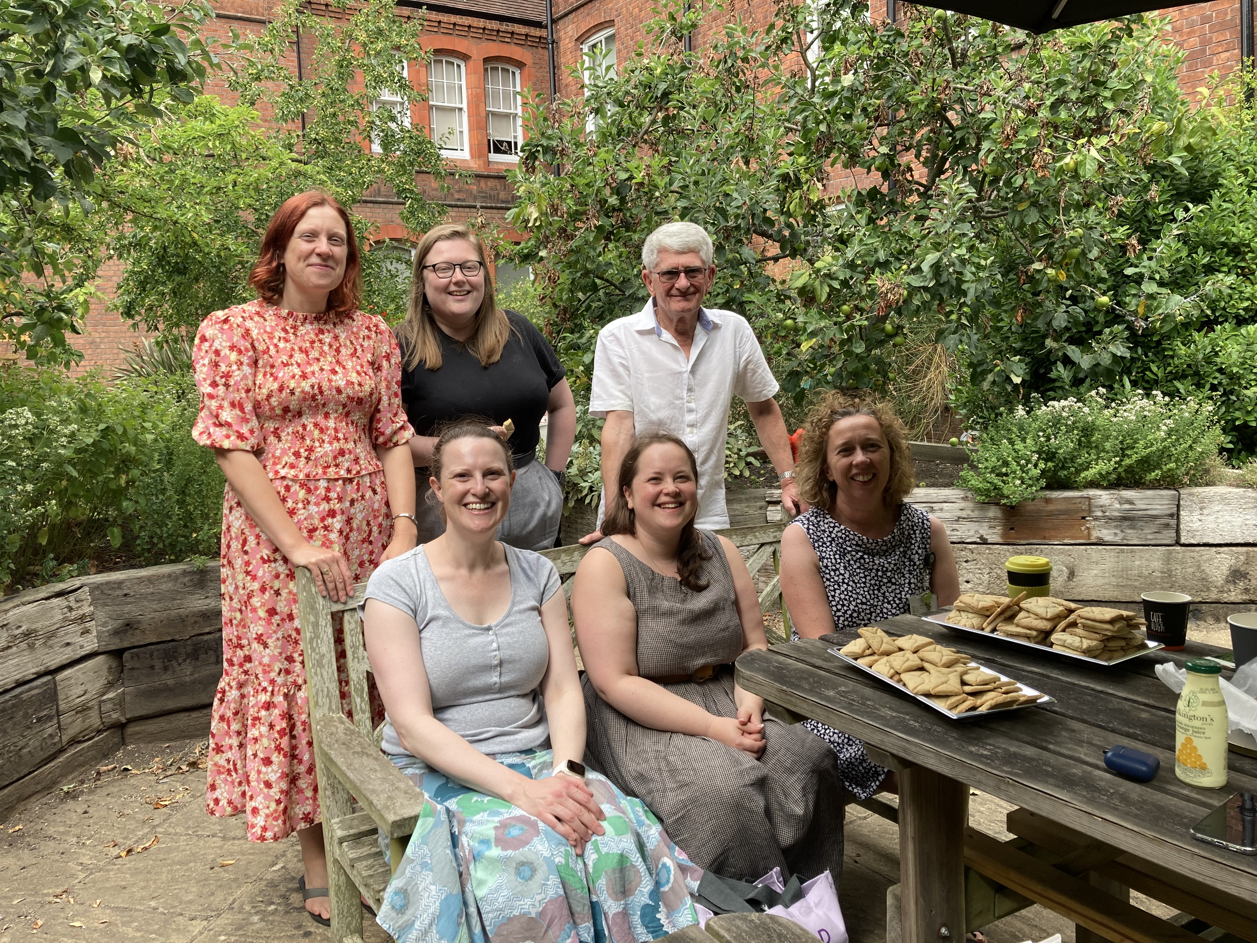 Image of volunteers and staff at a small garden party celebrating the first 100 transcriptions. Its shows people sitting by a table which has trays of biscuites on it
