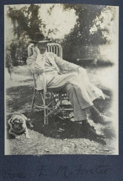 Man in a light-coloured suit reclining in a chair on a lawn. To his right, a pug is lying down.